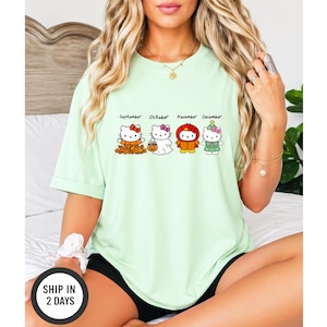 Puede incluir: Una camiseta de color verde claro con cuatro personajes de Hello Kitty que representan los meses de septiembre, octubre, noviembre y diciembre. El gr&aacute;fico muestra a los personajes con trajes de temporada, incluyendo hojas de oto&ntilde;o, un disfraz de fantasma, un pavo y un &aacute;rbol.