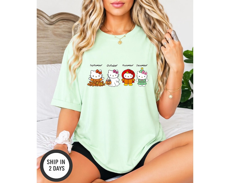 Puede incluir: Una camiseta de color verde claro con cuatro personajes de Hello Kitty que representan los meses de septiembre, octubre, noviembre y diciembre. El gr&aacute;fico muestra a los personajes con trajes de temporada, incluyendo hojas de oto&ntilde;o, un disfraz de fantasma, un pavo y un &aacute;rbol.