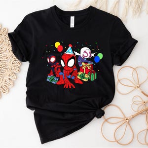 Puede incluir: Camiseta negra con una imagen de dibujos animados de Spider-Man, Spider-Gwen y otro personaje de Spider-Man celebrando un cumplea&ntilde;os con regalos y confeti.