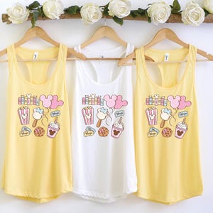 Pu&ograve; includere: Tre canottiere con colori giallo, bianco e giallo. Ogni canottiera presenta un design a cartoni animati con orecchie di Topolino, popcorn, gelato e una bevanda a forma di Topolino. Il testo "Magic" &egrave; stampato su ogni canottiera.