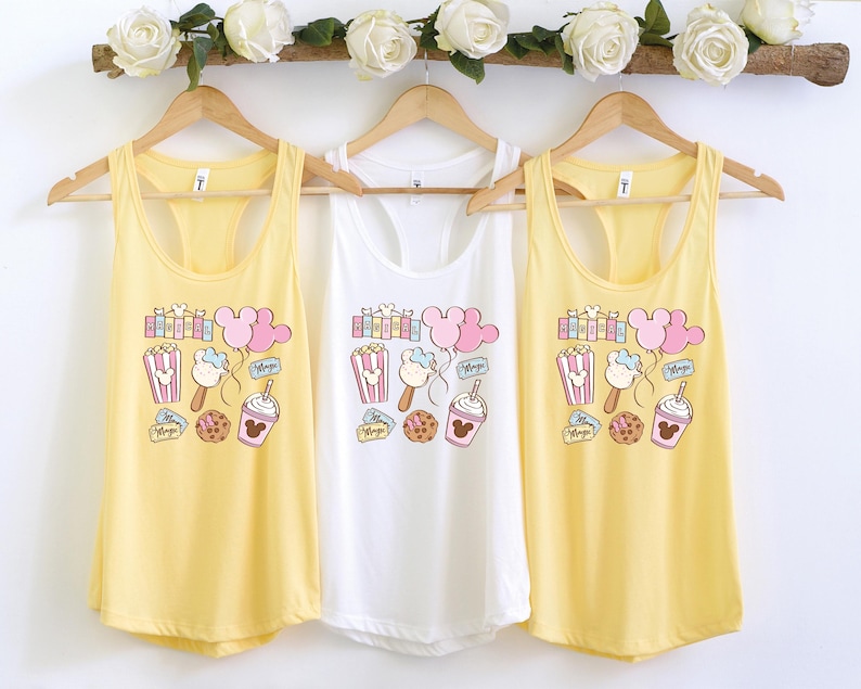 Pu&ograve; includere: Tre canottiere con colori giallo, bianco e giallo. Ogni canottiera presenta un design a cartoni animati con orecchie di Topolino, popcorn, gelato e una bevanda a forma di Topolino. Il testo "Magic" &egrave; stampato su ogni canottiera.