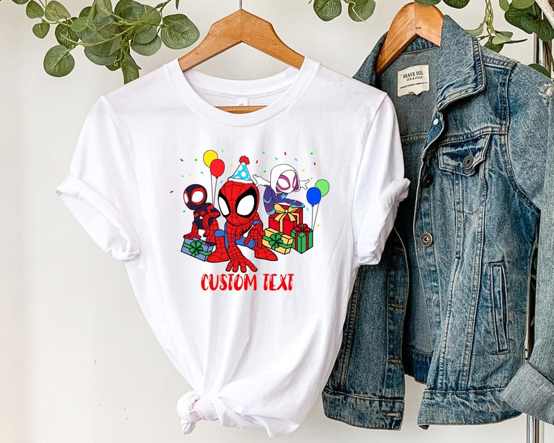 Puede incluir: Camiseta blanca con una imagen de dibujos animados de Spider-Man, Spider-Gwen y otro personaje de Spider-Man sosteniendo regalos y usando sombreros de fiesta. El texto "CUSTOM TEXT" est&aacute; impreso debajo de la imagen.