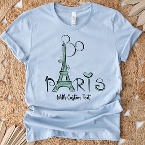 Op de afbeelding: Lichtblauw T-shirt met een groen ontwerp van de Eiffeltoren, hartjes en het woord "PARIS". De tekst "With Custom Text" staat onder het woord "PARIS". Het shirt is gemaakt van een zacht materiaal.