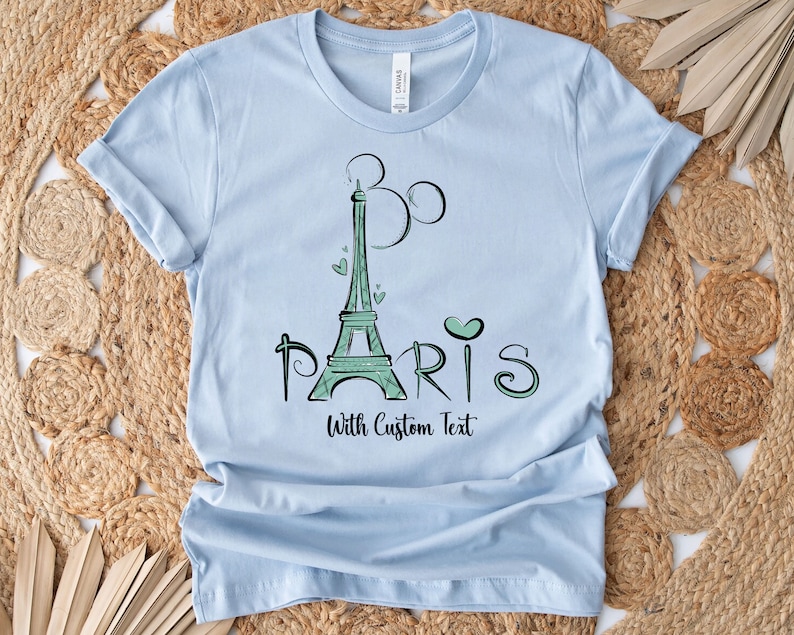 Op de afbeelding: Lichtblauw T-shirt met een groen ontwerp van de Eiffeltoren, hartjes en het woord "PARIS". De tekst "With Custom Text" staat onder het woord "PARIS". Het shirt is gemaakt van een zacht materiaal.
