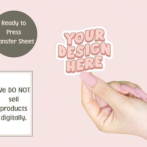 May include: A pink and white sticker with the text "YOUR DESIGN HERE" is held up by a hand with pink nails. The background is a light pink color. There is a dark green circle with the text "Ready to Press Transfer Sheet" and a white rectangle with the text "We DO NOT sell products digitally."