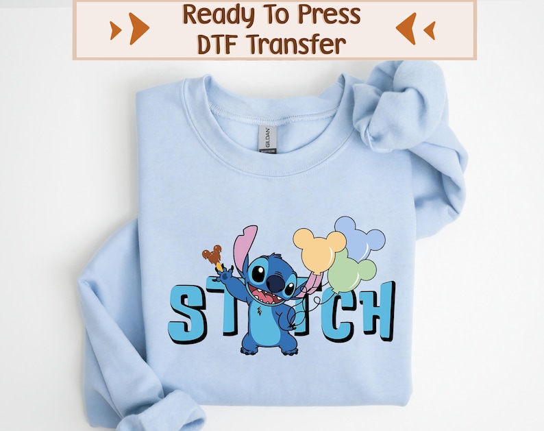 May include: Light blue sweatshirt featuring a graphic of the character Stitch holding balloons. The word "STITCH" is printed in large, blue letters below the character. The top of the image reads "Ready To Press DTF Transfer".