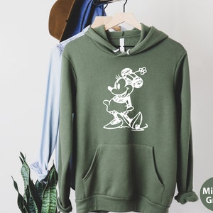 Puede incluir: Una sudadera con capucha verde con un contorno blanco de un rat&oacute;n de dibujos animados con un lazo. El texto "Military Green" est&aacute; en la esquina inferior derecha de la imagen.