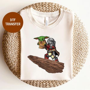 Puede incluir: Camiseta color crema con una transferencia DTF de un personaje al estilo Mandaloriano sosteniendo a un alienígena verde. El gráfico está sobre una formación rocosa marrón. El texto "DTF TRANSFER" está en un círculo marrón en la esquina superior izquierda.