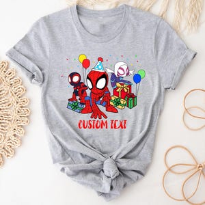 Puede incluir: Camiseta gris con una imagen de dibujos animados de tres personajes de Spider-Man celebrando un cumplea&ntilde;os con regalos y globos. El texto "CUSTOM TEXT" est&aacute; impreso debajo de la imagen.