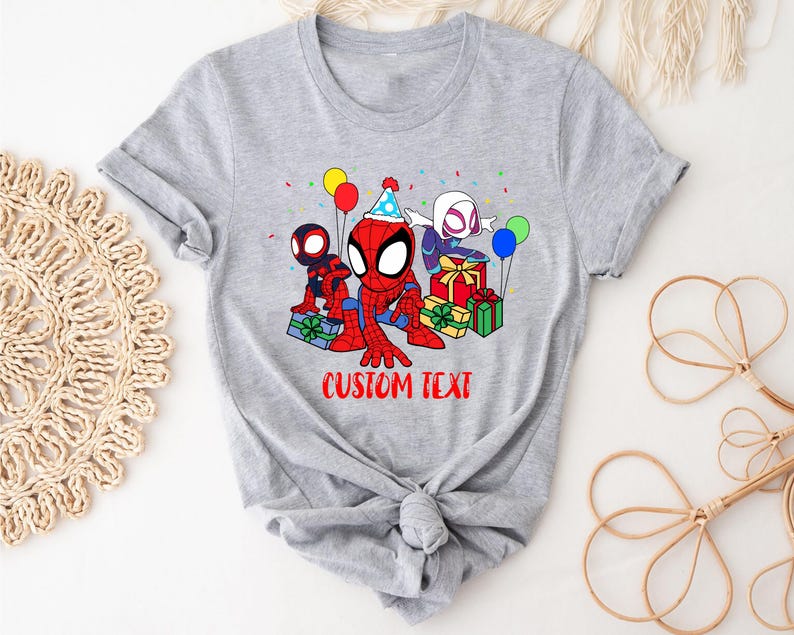 Puede incluir: Camiseta gris con una imagen de dibujos animados de tres personajes de Spider-Man celebrando un cumplea&ntilde;os con regalos y globos. El texto "CUSTOM TEXT" est&aacute; impreso debajo de la imagen.