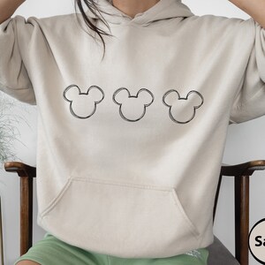 May include: A sand-coloured hoodie with a black outline of three Mickey Mouse heads on the front.