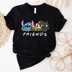 May include: Black t-shirt featuring a graphic of Stitch, Toothless, Grogu, and other characters. The word "FRIENDS" is printed in white block letters below the characters. The shirt is made of a soft material.