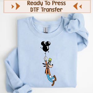 Peut inclure: Sweat-shirt bleu clair avec une image de dessin animé en noir et blanc de Dingo tenant un ballon en forme de Mickey Mouse. Le texte "Ready To Press DTF Transfer" est imprimé sur un rectangle brun en haut de l'image.