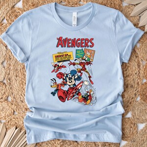 May include: Pale blue t-shirt with a vibrant graphic of Disney characters in an Avengers theme. The design includes Mickey Mouse, Donald Duck, and Iron Man, with the text "The Avengers" in red.