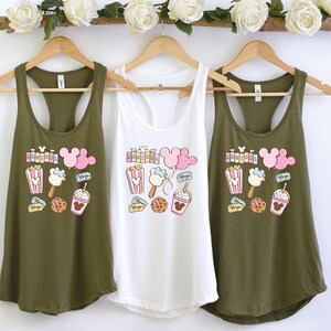Pu&ograve; includere: Tre canottiere verde oliva con una canottiere bianca al centro. Tutte e quattro le canottiere presentano una stampa grafica con orecchie di Topolino, popcorn, gelato e altri dolcetti del parco Disney.