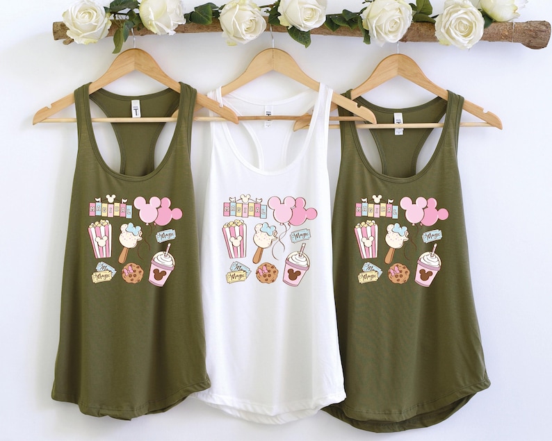 Pu&ograve; includere: Tre canottiere verde oliva con una canottiere bianca al centro. Tutte e quattro le canottiere presentano una stampa grafica con orecchie di Topolino, popcorn, gelato e altri dolcetti del parco Disney.