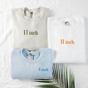 May include: Three sweatshirts in cream, white, and light blue are displayed. Each sweatshirt has the text "13 inch", "11 inch", and "4 inch" printed on them. The sweatshirts are folded and arranged on a white surface.