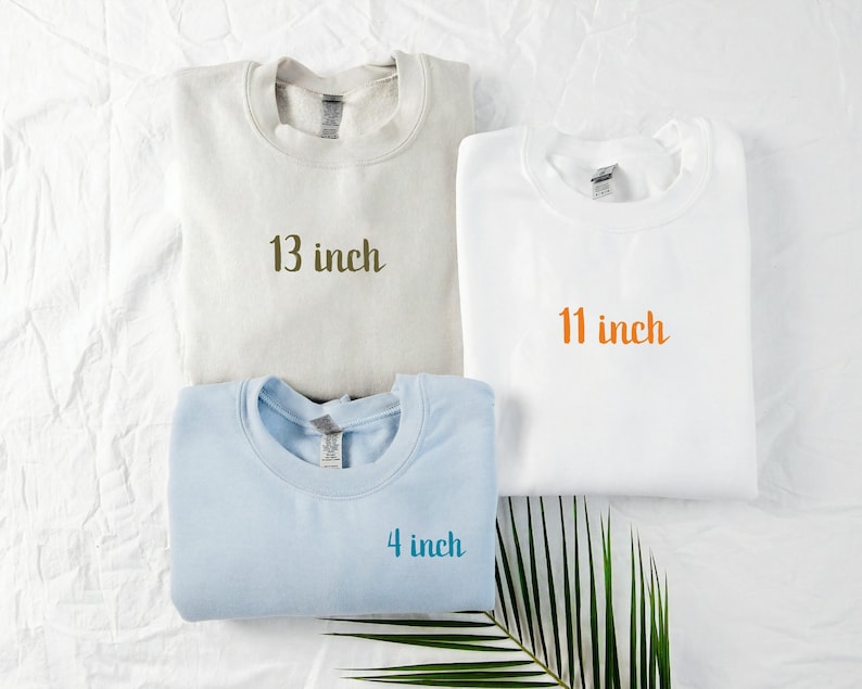 May include: Three sweatshirts in cream, white, and light blue are displayed. Each sweatshirt has the text "13 inch", "11 inch", and "4 inch" printed on them. The sweatshirts are folded and arranged on a white surface.