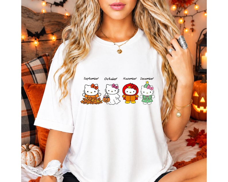Puede incluir: Una camiseta blanca con un gr&aacute;fico de cuatro personajes de Hello Kitty etiquetados como septiembre, octubre, noviembre y diciembre. Los personajes visten atuendos de temporada que incluyen hojas de oto&ntilde;o, un disfraz de fantasma y un &aacute;rbol.