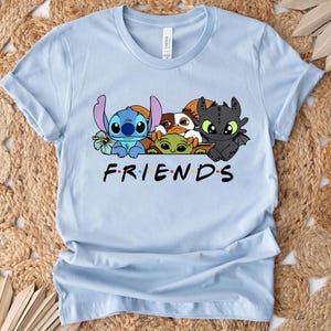 May include: Light blue t-shirt featuring a graphic of cartoon characters including Stitch, Baby Yoda, and Toothless. The word "FRIENDS" is printed in black with each letter in a different color. The shirt is made of a soft material.