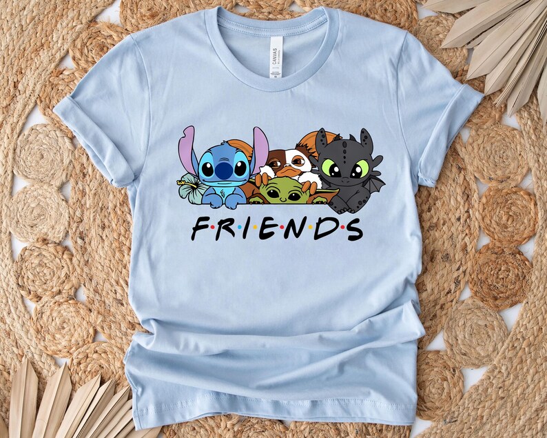 May include: Light blue t-shirt featuring a graphic of cartoon characters including Stitch, Baby Yoda, and Toothless. The word "FRIENDS" is printed in black with each letter in a different color. The shirt is made of a soft material.
