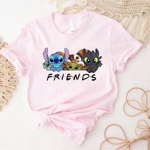 May include: A light pink t-shirt featuring a graphic of cartoon characters including Stitch, Baby Yoda, and Toothless. The word "FRIENDS" is printed in black block letters below the characters. The shirt is made of soft material.