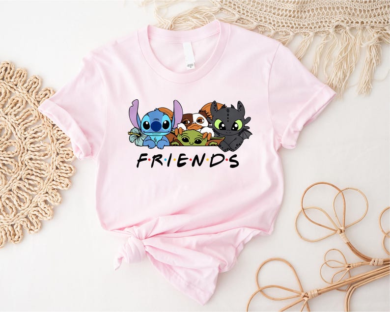 May include: A light pink t-shirt featuring a graphic of cartoon characters including Stitch, Baby Yoda, and Toothless. The word "FRIENDS" is printed in black block letters below the characters. The shirt is made of soft material.