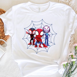 May include: White t-shirt with a blue and white spider web design featuring three cartoon Spider-Man characters. The characters are red, black, and white.