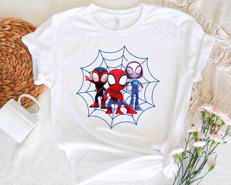 May include: White t-shirt with a blue and white spider web design featuring three cartoon Spider-Man characters. The characters are red, black, and white.