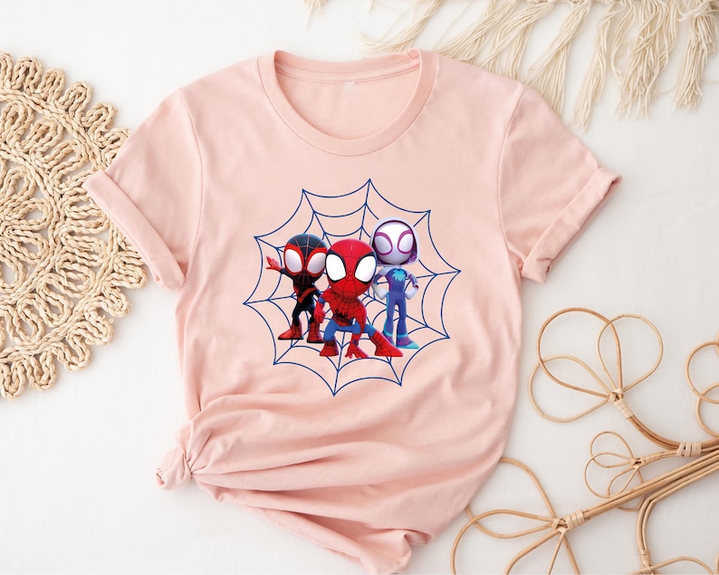 May include: A pink t-shirt with a graphic of three cartoon Spider-Man characters in a web. The characters are red, blue, and white.