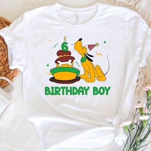 Könnte beinhalten: Weißes T-Shirt mit einem Cartoon-Hund, der eine Kerze auf einem Geburtstagskuchen ausbläst. Der Kuchen ist grün und gelb mit einem Knochen oben drauf. Der Text "BIRTHDAY BOY" ist unter dem Bild gedruckt.
