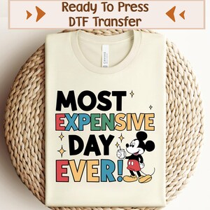 May include: A beige t-shirt featuring a colourful graphic design that reads "MOST EXPENSIVE DAY EVER!" with a cartoon image of Mickey Mouse holding a cup. The design is in a retro style with a rainbow colour scheme.