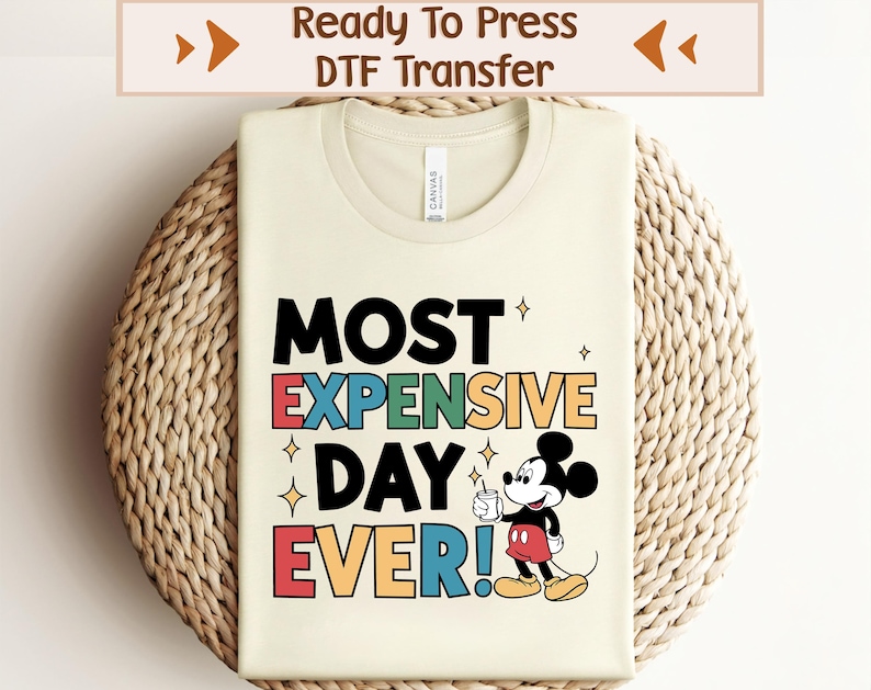 May include: A beige t-shirt featuring a colourful graphic design that reads "MOST EXPENSIVE DAY EVER!" with a cartoon image of Mickey Mouse holding a cup. The design is in a retro style with a rainbow colour scheme.