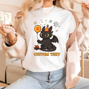 Puede incluir: Camiseta blanca con un dragón negro de dibujos animados que lleva un gorro de fiesta y sostiene un globo con el número 5. La camiseta tiene las palabras "CUSTOM TEXT" en letras naranjas.