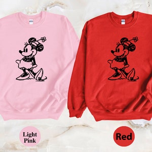 Puede incluir: Dos sudaderas con una imagen en blanco y negro de Minnie Mouse en la parte delantera. Una sudadera es rosa claro y la otra es roja.