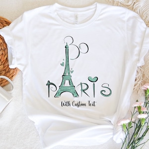 Op de afbeelding: Wit T-shirt met een turquoise Eiffeltoren ontwerp, Mickey Mouse oren en het woord "Paris". Het shirt bevat ook de tekst "With Custom Text" onder het woord "Paris".