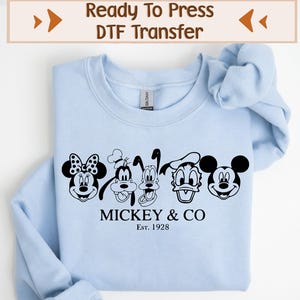 May include: Light blue sweatshirt with a black and white graphic of Mickey Mouse, Minnie Mouse, Donald Duck, Goofy, and Pluto. The graphic is accompanied by the text "Mickey & Co. Est. 1928".