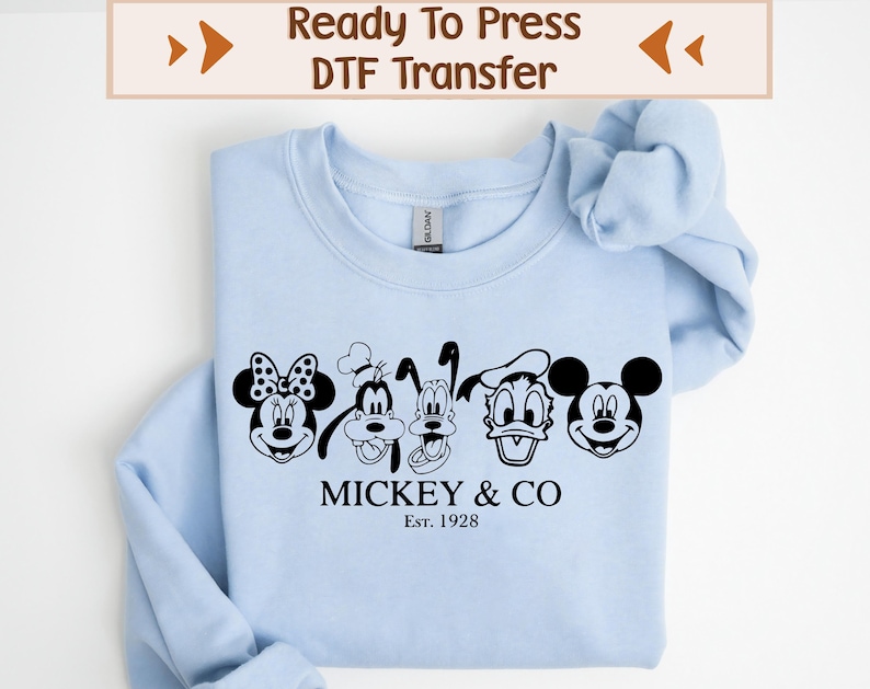 May include: Light blue sweatshirt with a black and white graphic of Mickey Mouse, Minnie Mouse, Donald Duck, Goofy, and Pluto. The graphic is accompanied by the text "Mickey & Co. Est. 1928".
