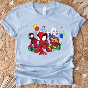 Puede incluir: Camiseta azul claro con una imagen de dibujos animados de tres personajes de Spider-Man celebrando un cumplea&ntilde;os. Los personajes llevan sombreros de fiesta y sostienen regalos. Hay globos y confeti en el fondo.