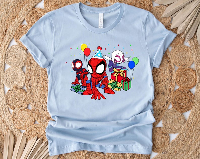 Puede incluir: Camiseta azul claro con una imagen de dibujos animados de tres personajes de Spider-Man celebrando un cumplea&ntilde;os. Los personajes llevan sombreros de fiesta y sostienen regalos. Hay globos y confeti en el fondo.