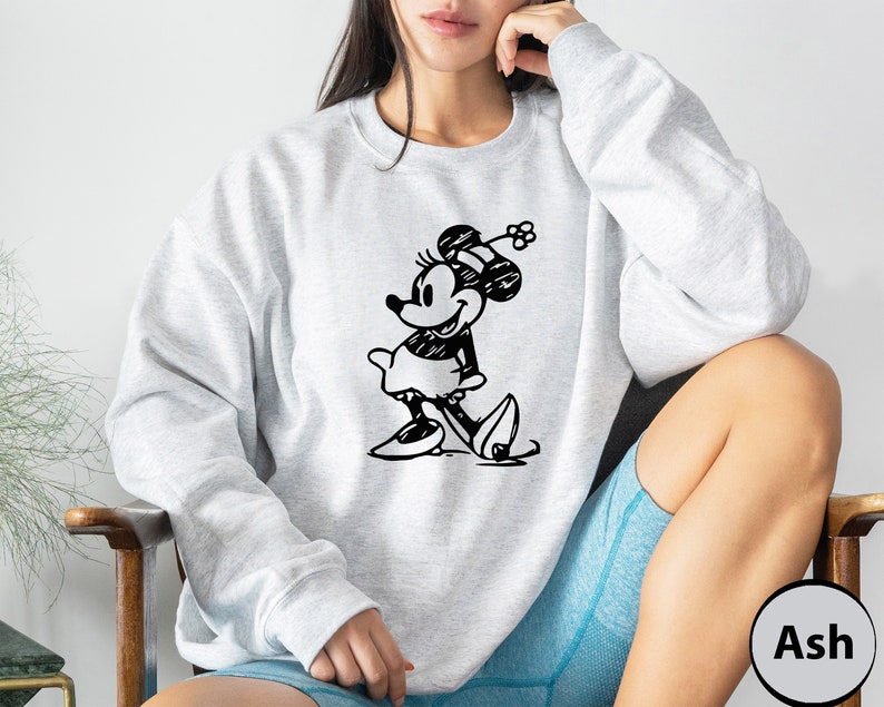 Puede incluir: Una sudadera gris claro con una ilustraci&oacute;n en blanco y negro de Minnie Mouse. La sudadera tiene cuello redondo y mangas largas. El texto "Ash" es visible en la esquina inferior derecha de la imagen.