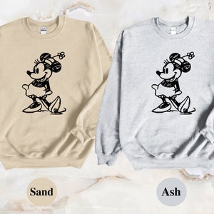 Puede incluir: Dos sudaderas con una ilustraci&oacute;n en blanco y negro de Mickey Mouse. La sudadera de la izquierda es de color arena y la sudadera de la derecha es de color ceniza.