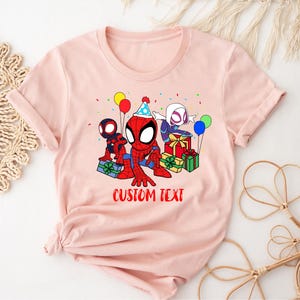 Puede incluir: Camiseta rosa con una imagen de dibujos animados de Spider-Man, Spider-Gwen y otro personaje de Spider-Man. Los personajes sostienen regalos y globos. El texto "CUSTOM TEXT" est&aacute; impreso debajo de la imagen.