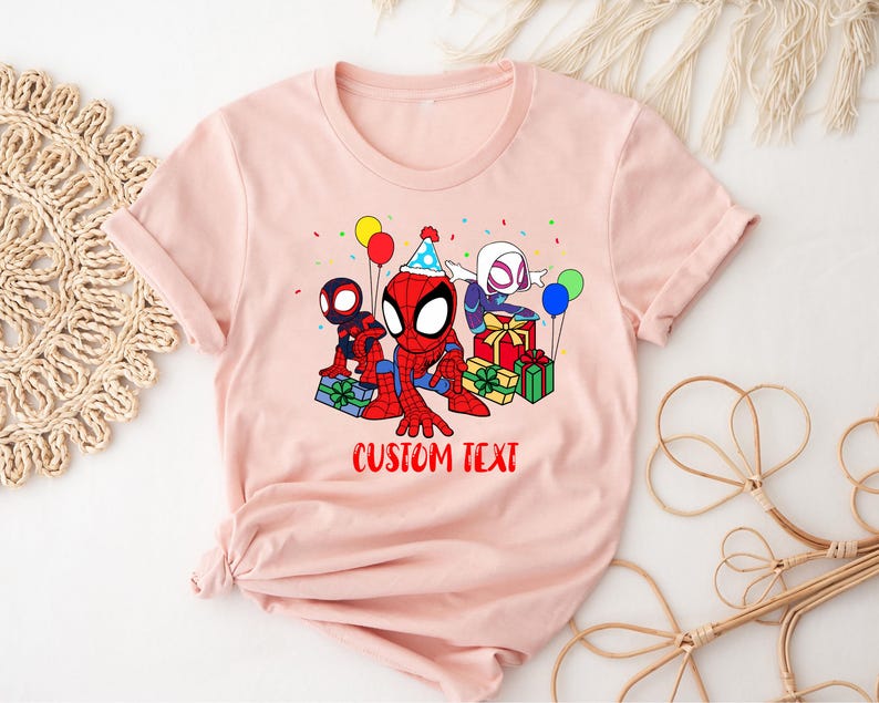 Puede incluir: Camiseta rosa con una imagen de dibujos animados de Spider-Man, Spider-Gwen y otro personaje de Spider-Man. Los personajes sostienen regalos y globos. El texto "CUSTOM TEXT" est&aacute; impreso debajo de la imagen.