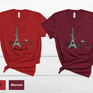 Op de afbeelding: Twee T-shirts, &eacute;&eacute;n rood en &eacute;&eacute;n bordeauxrood, elk met een turquoise Eiffeltoren-afbeelding en het woord "Paris" en de tekst "With Custom Text". De shirts worden gepresenteerd op een wit houten oppervlak.