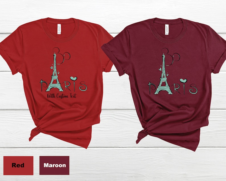 Op de afbeelding: Twee T-shirts, &eacute;&eacute;n rood en &eacute;&eacute;n bordeauxrood, elk met een turquoise Eiffeltoren-afbeelding en het woord "Paris" en de tekst "With Custom Text". De shirts worden gepresenteerd op een wit houten oppervlak.