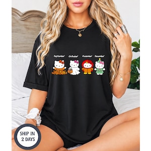 Puede incluir: Una camiseta negra de manga corta con un estampado gr&aacute;fico de cuatro personajes de Hello Kitty etiquetados como September, October, November y December. Los personajes visten trajes de temporada que representan hojas de oto&ntilde;o, un fantasma, un pavo y un &aacute;rbol.