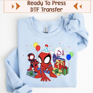 以下が含まれることがあります： 薄い青色のスウェットシャツに、DTF転写のデザインが施されています。3人のスパイダーマンの漫画キャラクターが、プレゼントと風船を持って誕生日を祝っています。画像の上部には「Ready To Press DTF Transfer」というテキストが表示されています。