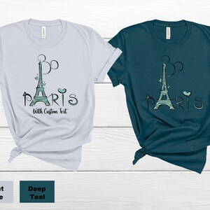 Op de afbeelding: Twee t-shirts, &eacute;&eacute;n lichtblauw en &eacute;&eacute;n diepblauwgroen, met een grafische afbeelding van de Eiffeltoren in zeegroen en het woord "Paris". Het ontwerp bevat hartjes en een op Mickey Mouse ge&iuml;nspireerd ontwerp. De tekst "With Custom Text" is ook inbegrepen.