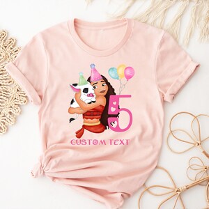 Pu&ograve; includere: Maglietta rosa con un'immagine di cartone animato di una ragazza con i capelli castani e un maiale. La ragazza indossa un vestito rosa e rosso e tiene un maiale. Il numero 5 &egrave; in rosa e il testo "CUSTOM TEXT" &egrave; sotto l'immagine.